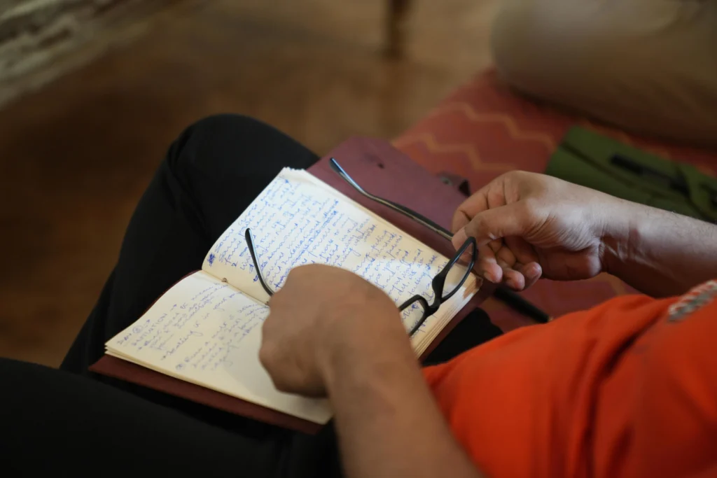 Close-up of a man writing in a notebook, glasses in hand — reflecting during a retreat.