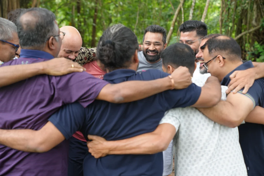 Men standing in a circle with arms around each other, sharing a joyful moment of connection.